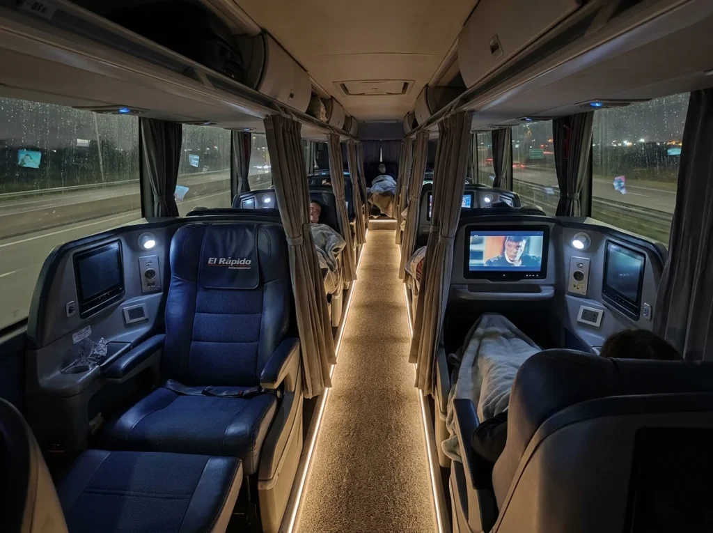 The interior of a modern, long-distance bus in Argentina at night. The seats are wide, fully-reclining “cama” seats with personal entertainment screens and curtains for privacy. The aisle is softly lit, conveying comfort and practicality for overnight journeys