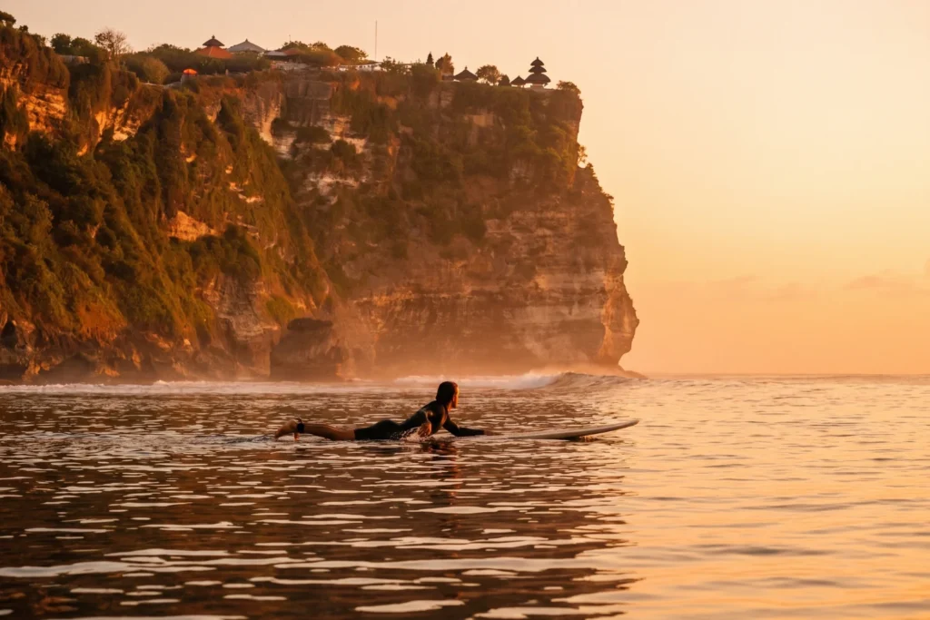 surfer paddling out at Uluwatu break at dawn, glassy water, cliff face in background, no other surfers visible, warm orange light