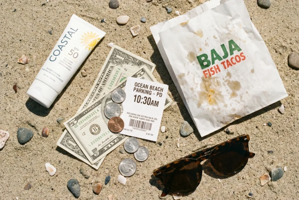 flat-lay style shot of beach trip essentials on sand — sunscreen, cash and coins, parking stub, a fish taco wrapper, sunglasses, all arranged casually, bright midday light