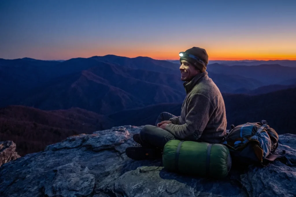 A backpacker sitting on a rocky summit at dusk, headlamp on, sleeping bag rolled beside them | Triumphant, serene | Twilight blue-purple with last orange on the horizon | Indigo, slate, amber