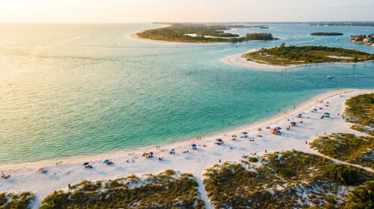 aerial view of a curving Florida Gulf Coast shoreline at golden hour, turquoise shallow water meeting white sand, scattered beachgoers with colorful umbrellas, sea oats framing the foreground