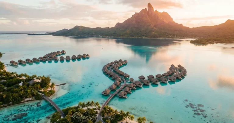 Aerial view of Bora Bora's turquoise lagoon at golden hour, the smell of salt and frangipani drifting up from the water, Mount Otemanu rising sharply in the background, overwater bungalows casting long shadows across the glassy surface, shot from a high angle with warm light filtering through thin clouds